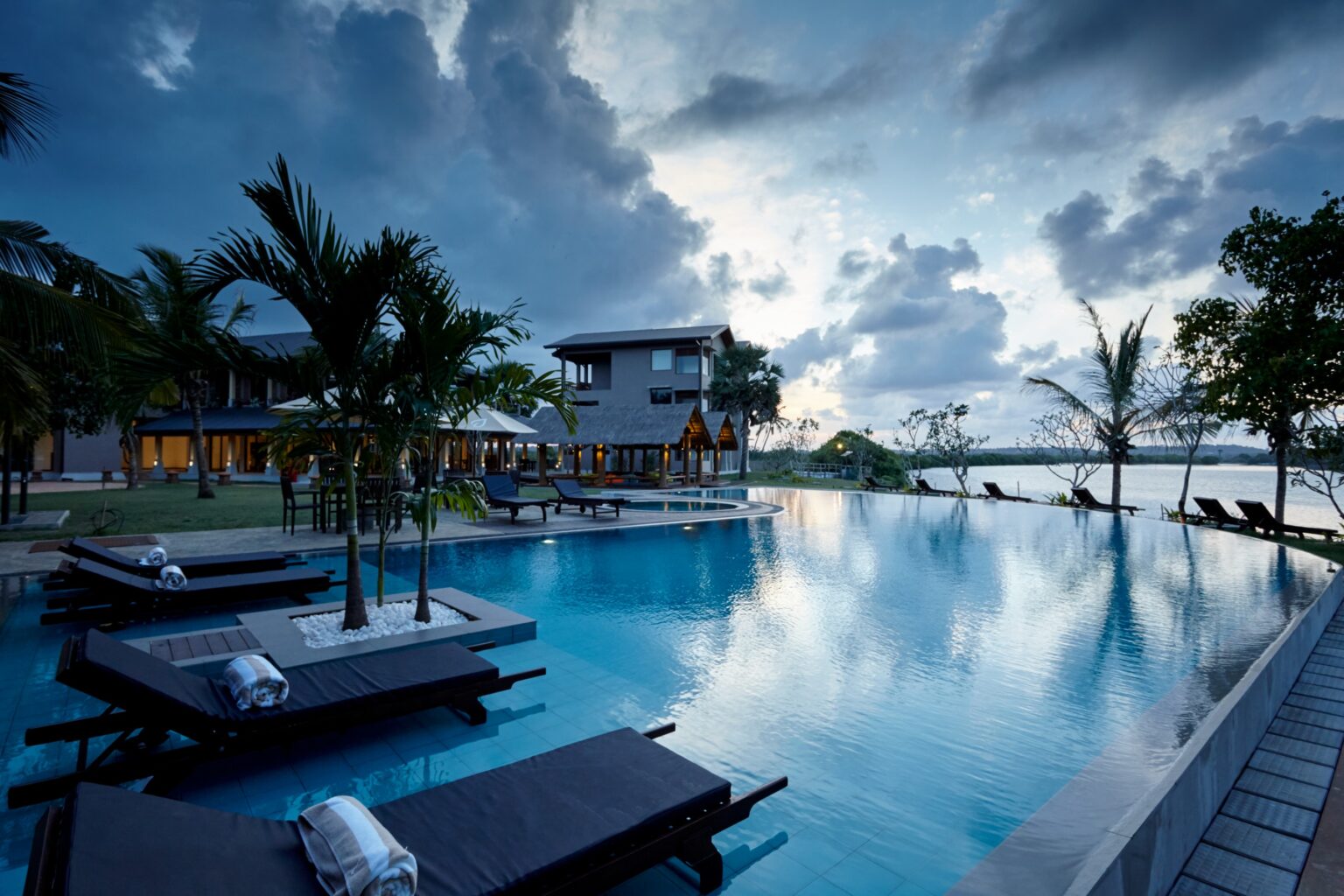 An infinity pool with sunbeds and palms, running out towards the lagoon under a cloudy sky
